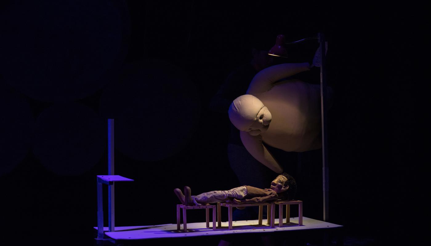 A small puppet lays on a bench while a large, puffy, white puppet hovers above