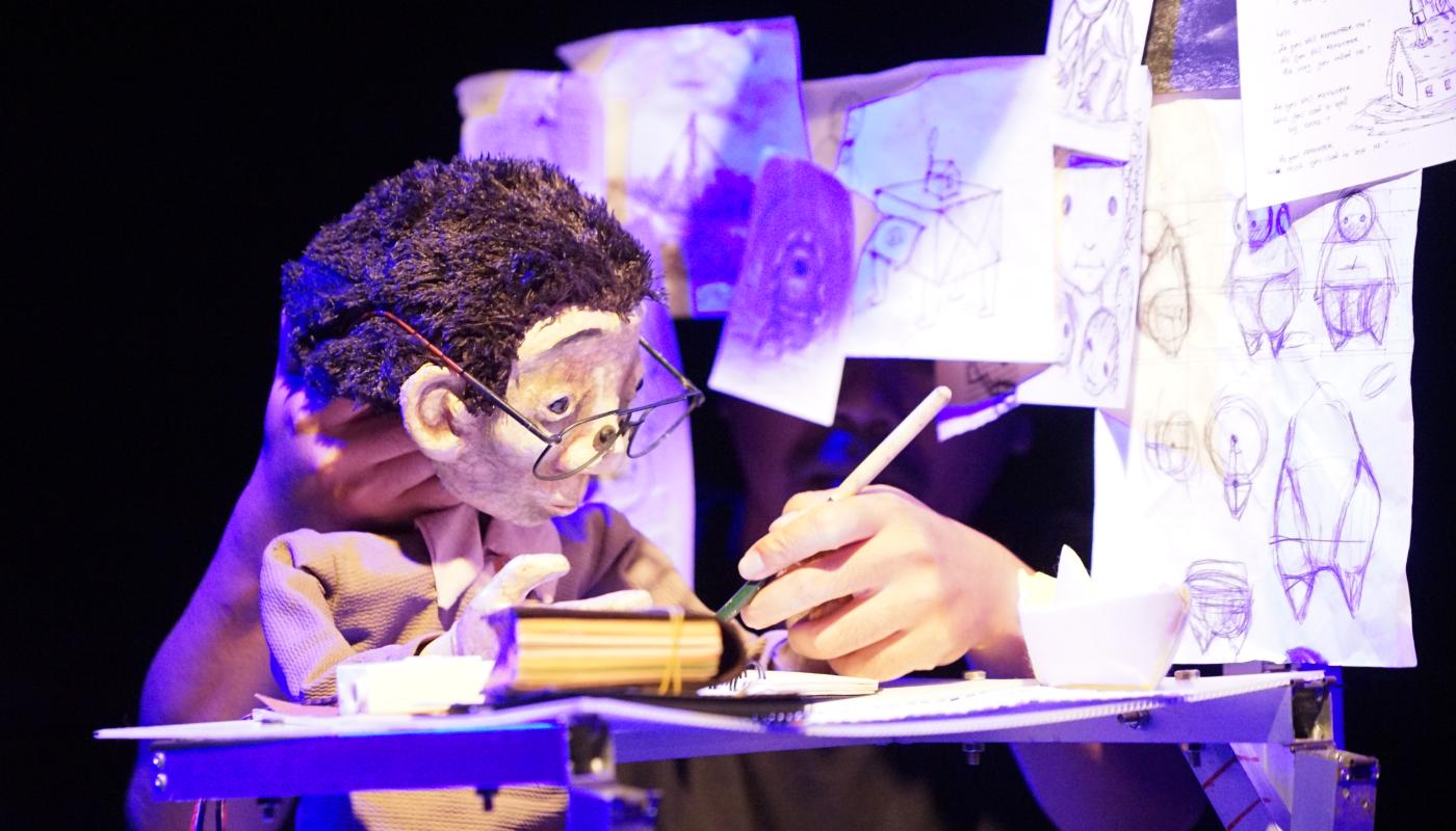 A puppet sits at a desk with drawings around them, you can see the puppeteers hand moving the head of the puppet