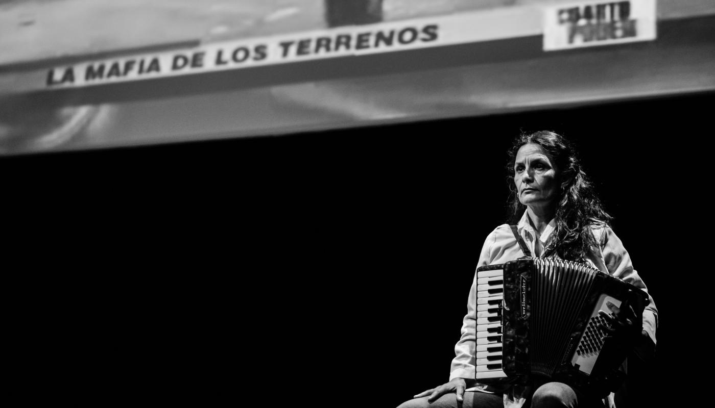 Black and white photo of a woman holding an accordion, sitting on stage in front of a projected image
