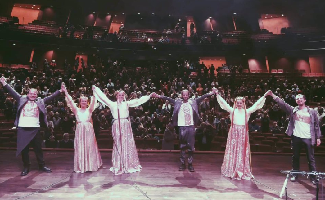 Three men and three women stand on a stage with their hands in the air in front of a full audience