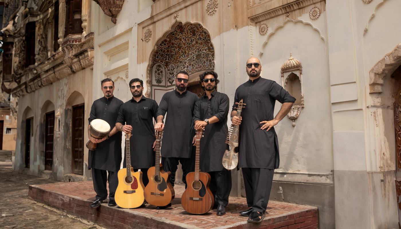 Khumariyaan by Bilal Khan Five men in traditional black outfits standing together with various instruments in front of a historic building in Pakistan