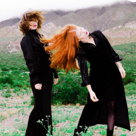 Femina by Inti Patron 2 Two women in black, one with dark hair and one with red hair, shake their heads in motion in front of a green landscape.