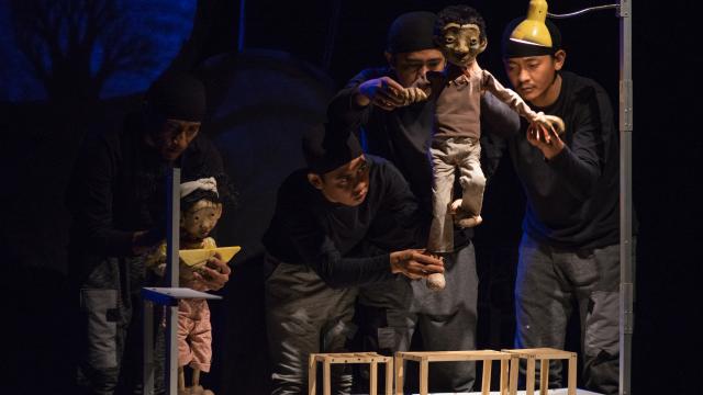Three puppeteers move a puppet on a small stage with a yellow light above them