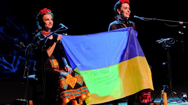 Kurbasy, courtesy of the band Two women sit on stage with microphones holding a Ukrainian flag between them