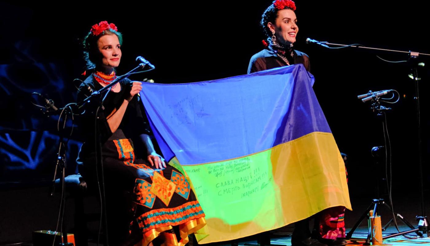 Kurbasy, courtesy of the band Two women sit on stage with microphones holding a Ukrainian flag between them