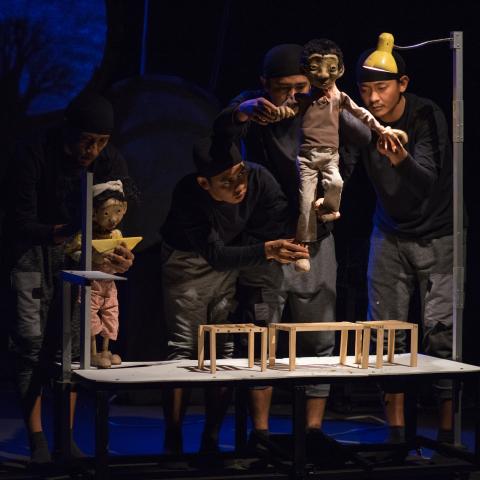 Three puppeteers move a puppet on a small stage with a yellow light above them