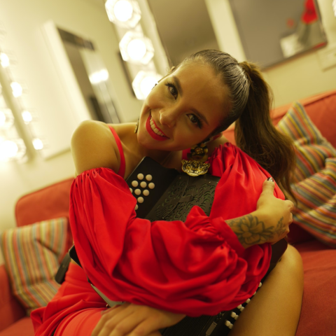 A woman in a red dress smiles while hugging an accordion in a backstage setting