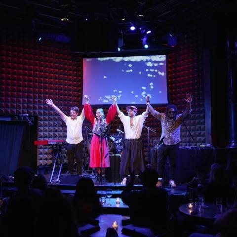 Four musicians (three male, one female), join hands in the air on stage while the audience claps.
