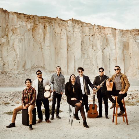 Seven people standing together with various instruments in front of a large white cliff